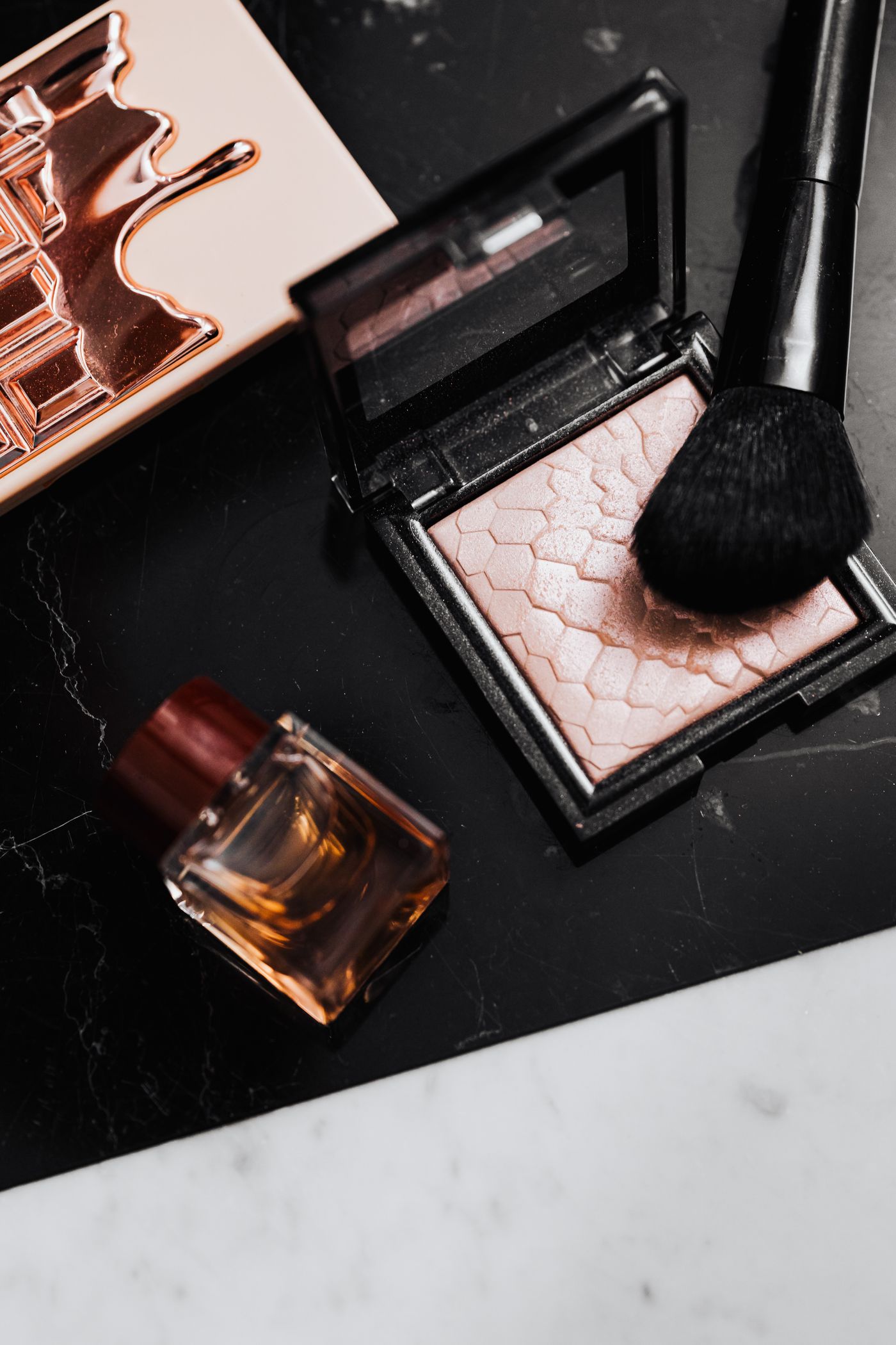 A flat lay of luxury makeup products, including a highlighter, brush and perfume bottle on a marble surface